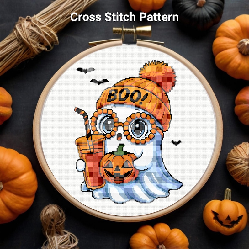 May include: Cross-stitch pattern featuring a cute ghost wearing an orange beanie with "BOO!" on it, round glasses, and holding a pumpkin and a drink. The design is set in a wooden embroidery hoop, with pumpkins and bats around it.
