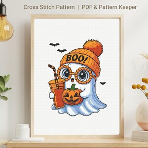 May include: Cross-stitch pattern of a cartoon ghost wearing an orange beanie with the word "BOO!" and glasses, holding a pumpkin and a drink. The ghost is light blue with a white face. The image is framed and includes the text "Cross Stitch Pattern | PDF & Pattern Keeper".