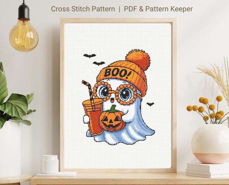 May include: Cross-stitch pattern of a cartoon ghost wearing an orange beanie with the word "BOO!" and glasses, holding a pumpkin and a drink. The ghost is light blue with a white face. The image is framed and includes the text "Cross Stitch Pattern | PDF & Pattern Keeper".