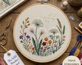 Op de afbeelding: Een ronde houten borduurring met een stoffen paneel voorzien van een handgeborduurd bloemmotief met witte paardenbloemen, groen gras en kleine kleurrijke wilde bloemen. Een wit label vermeldt de tekst The Wild Meadow Project.