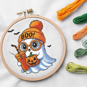 May include: Cross-stitch embroidery of a cartoon ghost wearing an orange beanie and glasses, holding a pumpkin and a drink. The word "BOO!" is on the hat. Includes orange, green, and yellow embroidery floss. The design is set in a wooden hoop.