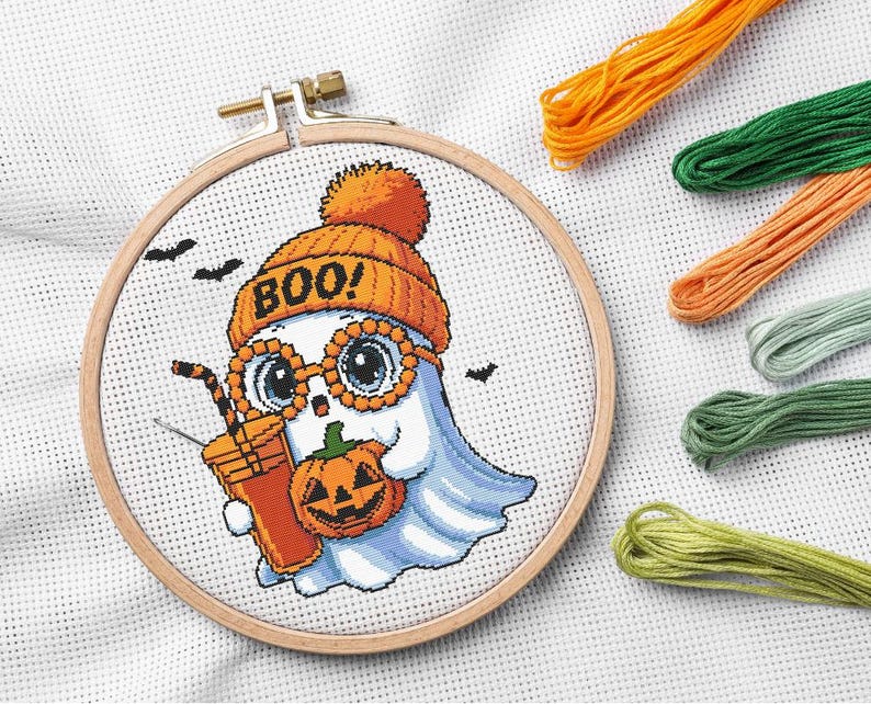 May include: Cross-stitch embroidery of a cartoon ghost wearing an orange beanie and glasses, holding a pumpkin and a drink. The word "BOO!" is on the hat. Includes orange, green, and yellow embroidery floss. The design is set in a wooden hoop.