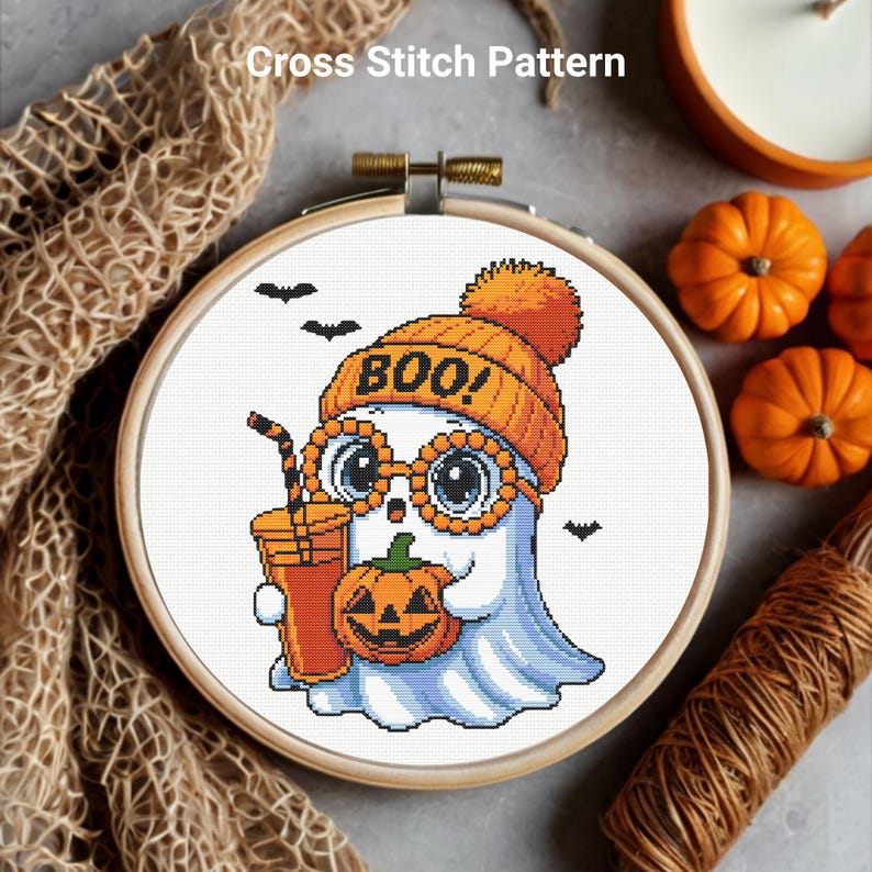 May include: A cross-stitch pattern in a wooden hoop features a cartoon ghost wearing an orange beanie and glasses, holding a pumpkin and a drink. The word "BOO!" is on the hat. Small orange pumpkins and bats are in the background.