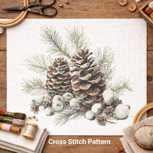 May include: Cross-stitch pattern featuring two detailed brown pine cones with green pine needles and white accents. The text "Cross Stitch Pattern" is at the bottom. The design is on white fabric, with embroidery supplies in the background.