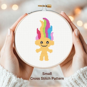 May include: A cross stitch pattern of a yellow troll with rainbow hair. The pattern is in a wooden embroidery hoop and is labeled "Small Cross Stitch Pattern".