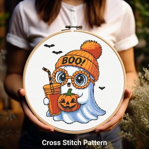 May include: A cross-stitch pattern featuring a cartoon ghost wearing an orange beanie with "BOO!" on it and round glasses. The ghost holds a pumpkin and a drink. The design is set in a wooden embroidery hoop, with the text "Cross Stitch Pattern" at the bottom.