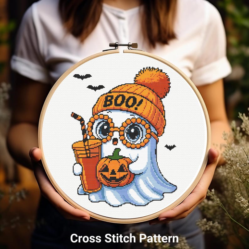 May include: A cross-stitch pattern featuring a cartoon ghost wearing an orange beanie with "BOO!" on it and round glasses. The ghost holds a pumpkin and a drink. The design is set in a wooden embroidery hoop, with the text "Cross Stitch Pattern" at the bottom.