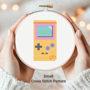 May include: A cross stitch pattern of a yellow handheld video game console with a purple and green screen. The pattern is displayed in a wooden embroidery hoop.