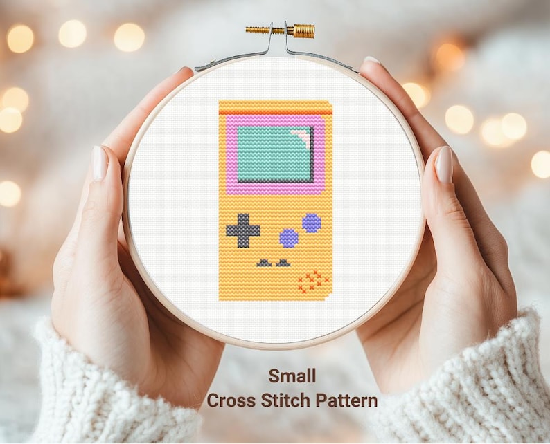 May include: A cross stitch pattern of a yellow handheld video game console with a purple and green screen. The pattern is displayed in a wooden embroidery hoop.
