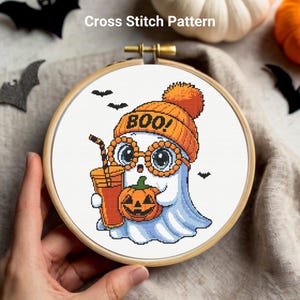 May include: Cross stitch pattern featuring a cartoon ghost wearing an orange beanie with "BOO!" on it and glasses. The ghost holds a pumpkin and a drink. The design is set in a wooden embroidery hoop.