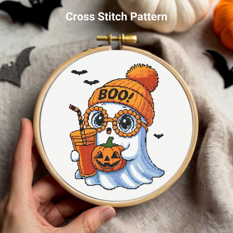 May include: Cross stitch pattern featuring a cartoon ghost wearing an orange beanie with "BOO!" on it and glasses. The ghost holds a pumpkin and a drink. The design is set in a wooden embroidery hoop.