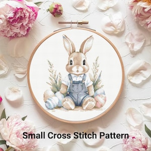 Könnte beinhalten: Ein Kreuzstichmuster mit einem niedlichen Hasen, der eine Latzhose trägt und Ostereier hält. Der Hase ist von Blumen und Grün umgeben. Das Muster ist perfekt für Anfänger und wird Ihrem Zuhause einen Hauch von Frühling verleihen.