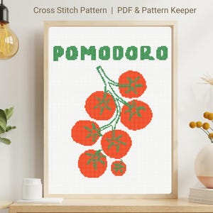 May include: A framed cross-stitch pattern featuring the word "POMODORO" in green above a cluster of red tomatoes with green stems and leaves. The artwork is displayed on a white background.