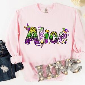 May include: Pink sweatshirt with the name "Alice" in a colorful, glittery font with Mardi Gras themed decorations.