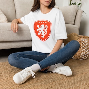 Könnte beinhalten: Weißes T-Shirt mit rotem Schild, das ein weißes Löwenemblem und den Text "KNVB" zeigt. Das Shirt besteht aus weichem Material und eignet sich für Freizeitkleidung. Das Design ist mittig auf der Vorderseite des Shirts platziert.