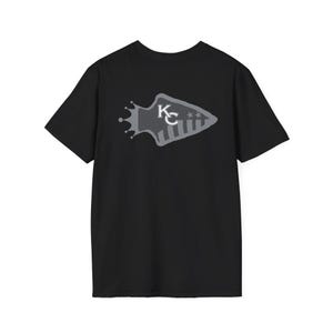May include: Black t-shirt featuring a gray arrowhead design with a crown and the letters "KC". The arrowhead has stripes and two stars. The shirt is a short-sleeved crew neck.