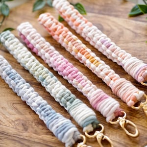 May include: Five colourful macrame keychains with gold key rings. The keychains are made with a variety of pastel colours, including pink, blue, green, and yellow.