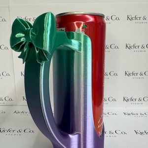 May include: A 3D-printed mug with a handle and a bow, in a gradient of green, blue, and purple. A red can is inside the mug. The mug is designed to hold a standard-sized beverage can. The background features a repeating logo.
