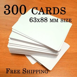 Puede incluir: Un montón de 300 tarjetas blancas, cada una de 6,3 x 8,8 cm. Las tarjetas son rectangulares con esquinas redondeadas. El texto "300 CARDS" y "63x88 MM SIZE" está en negro. Las tarjetas están apiladas sobre una superficie de madera.