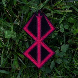 May include: A black and red patch featuring a stylized, symmetrical design. The patch has a black center with a red outline and is set against a backdrop of green grass.