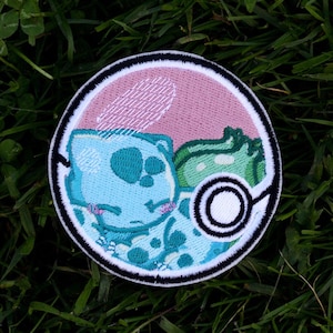 Op de afbeelding: Een ronde geborduurde patch met een blauw en groen cartoonfiguur in een roze en witte cirkel. De patch heeft een zwarte rand en ligt op groen gras.