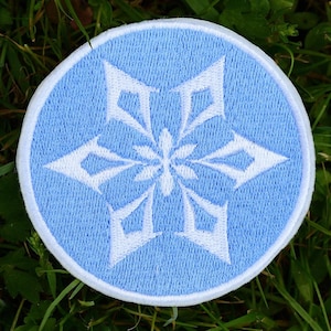 May include: A round, light blue patch with a white snowflake design. The snowflake has six points and is centered on the patch. The patch has a white border and is set against a background of green grass.
