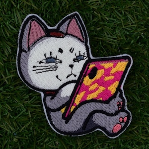 Peut inclure: Un chat gris et blanc avec des oreilles roses regarde un téléphone rose et jaune avec un écran noir.