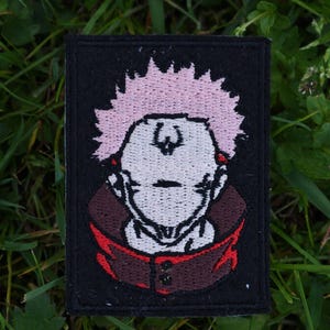 May include: Embroidered patch featuring a cartoon character with pink hair, a red and black shirt, and a white face. The patch is on a black background.