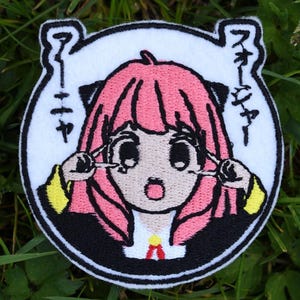 以下が含まれることがあります： ピンクの髪、黒い目、黒と黄色の服を着たアニメの女の子が描かれた白い丸い刺繍パッチ。女の子は手で目を大きく開けています。パッチには「アイヤー」という日本語が書かれています。
