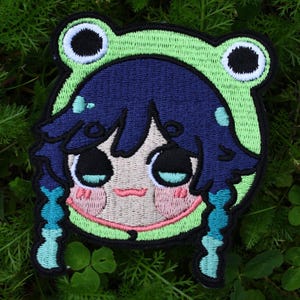 Könnte beinhalten: Gestickter Aufnäher mit einer Comicfigur mit blauen Haaren, die einen grünen Froschhut trägt. Die Figur hat ein rosafarbenes Gesicht und blaue Augen.