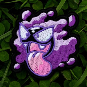 Könnte beinhalten: Gestickter Aufnäher mit einem lila Cartoon-Geist mit schwarzer Umrandung. Der Geist hat weiße Augen, eine rosa Zunge und ein breites Grinsen. Der Aufnäher befindet sich vor einem Hintergrund aus grünem Klee.