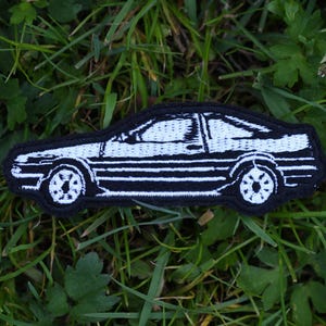 Könnte beinhalten: Weißer gestickter Aufnäher mit einem Auto auf schwarzem Hintergrund. Das Auto ist ein klassischer japanischer Sportwagen mit weißem Umriss und schwarzen Details.