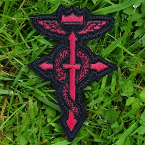 Puede incluir: Parche bordado con un diseño estilizado en rojo y negro. El parche tiene una forma de cruz central con un elemento serpentino y una parte superior en forma de corona. El parche está sobre un fondo de hierba verde.