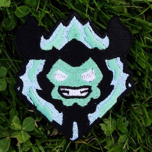 May include: A black and green embroidered patch featuring a stylized monster face. The monster has a green face, white teeth, and light blue accents. The patch is set against a background of green grass and clover.