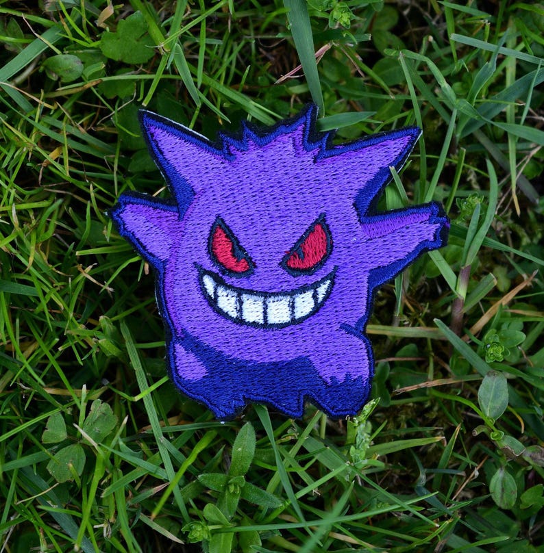 Op de afbeelding: Paarse en blauwe geborduurde patch van een cartoon-spook met rode ogen en een brede grijns. Het spook bevindt zich op een groene gras achtergrond.