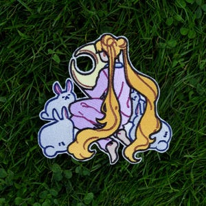 Könnte beinhalten: Gestickter Aufnäher mit einer Zeichentrickfigur mit langen blonden Haaren und einem rosa Outfit, umgeben von hellblauen Kaninchen. Der Aufnäher hat einen weißen Rand, vor einem grünen Hintergrund.