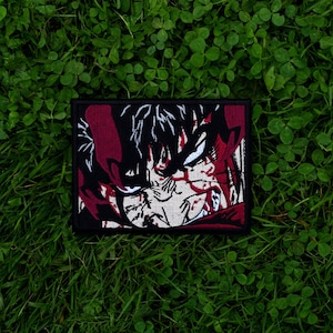 May include: A rectangular patch featuring a detailed anime-style illustration. The design showcases a character with white hair and a determined expression, set against a black background with red accents. The patch is bordered in black.