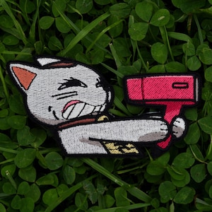 Pode incluir: Patch bordado com um gato de desenho animado segurando um objeto rosa. O gato é branco com detalhes marrons e pretos, e o objeto tem uma forma retangular com uma linha preta. O patch está em um fundo verde.