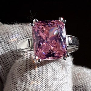 Może przedstawiać: Srebrny pierścionek z dużym, kwadratowym, różowym kamieniem szlachetnym. Kamień szlachetny jest fasetowany i osadzony w szynie.