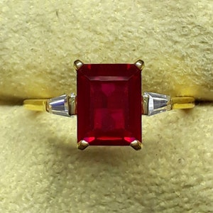 Puede incluir: Un anillo de oro con una gran piedra de rubí de talla esmeralda y dos diamantes más pequeños de talla baguette a cada lado.