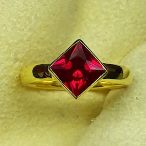 Może przedstawiać: Złoty pierścionek z kwadratowym, fasetowanym, jasno czerwoną kamieniem szlachetnym. Kamień jest osadzony w złotej oprawie, a obrączka jest z gładkiego, polerowanego złota. Pierścionek jest prezentowany na jasnożółtej powierzchni.