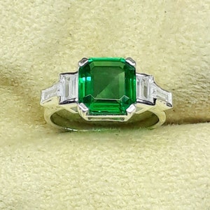 May include: A silver ring featuring a large emerald green gemstone flanked by two smaller baguette-cut diamonds.