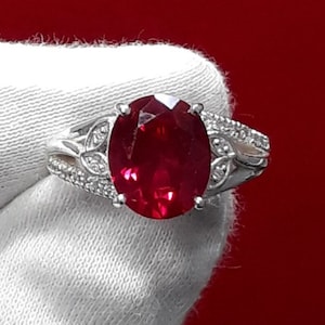 Peut inclure: Une bague en argent ornée d'une grosse pierre rouge foncé de taille ovale. Le corps de la bague est divisé et orné de petites pierres claires et de motifs en forme de feuilles, sur fond rouge vif.