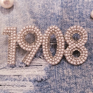 May include: A silver-toned brooch shaped like the year 1908, made of white pearls. The brooch is on a blue denim background.