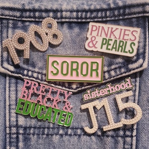May include: A collection of six enamel and pearl-embellished pins attached to a blue acid-wash denim fabric. The pins feature the text 1908, PINKIES & PEARLS, SOROR, PRETTY BLACK & EDUCATED, sisterhood, and J15 in pink, green, and white color schemes.