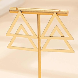 May include: Gold metal earring stand with two pairs of triangle shaped earrings. The stand has a round base and a single upright post.