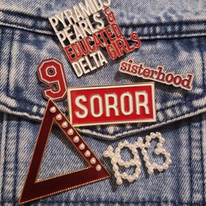 May include: A selection of enamel pins with diverse designs and text, such as "Pyramids Pearls & Educated Delta Girls", "Soror", "Sisterhood", a number 9, a triangle, and the number "1913". The pins are red, white, and gold.