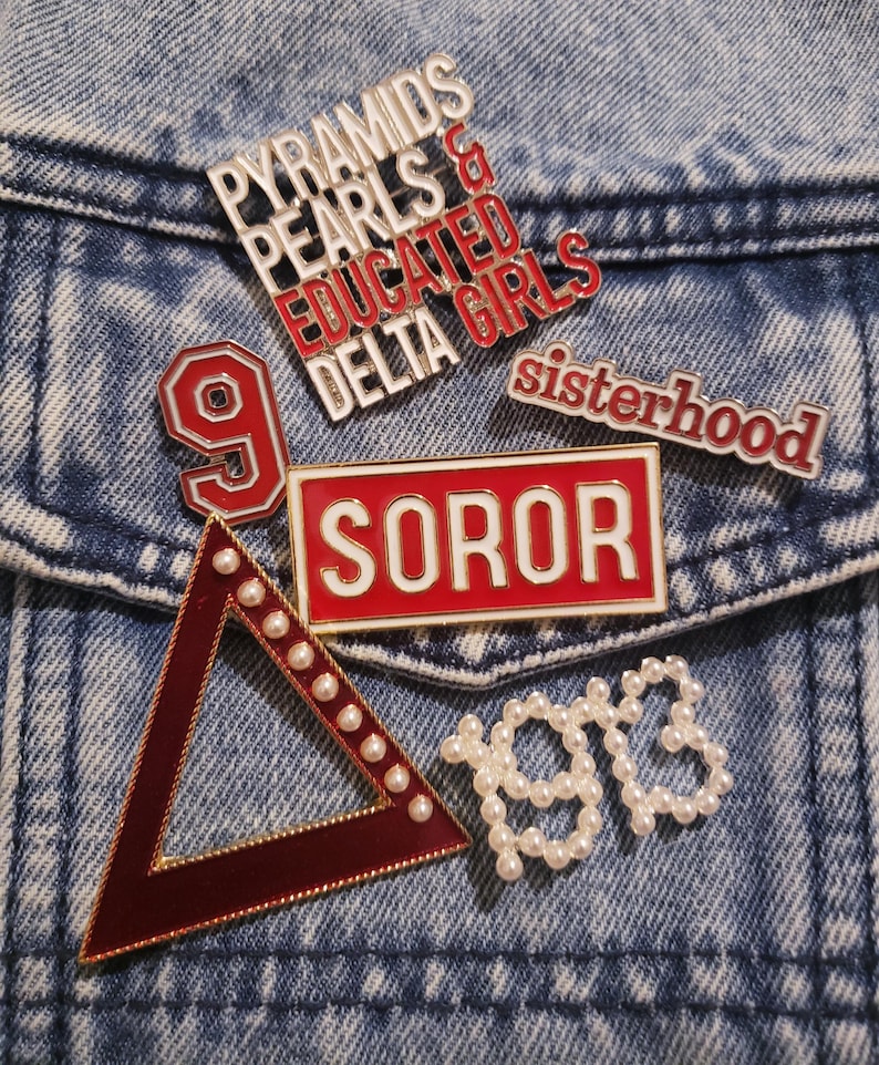May include: A selection of enamel pins with diverse designs and text, such as "Pyramids Pearls & Educated Delta Girls", "Soror", "Sisterhood", a number 9, a triangle, and the number "1913". The pins are red, white, and gold.