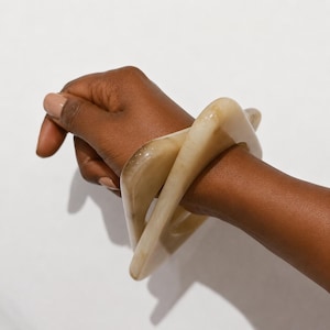 May include: A square-shaped bangle bracelet with rounded corners, crafted from a smooth, translucent material in a light beige and cream marbled pattern. The bracelet features a wide, thick band design that encircles the wrist with a polished finish.