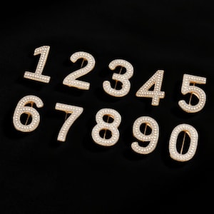 May include: A collection of gold-toned number pins, each encrusted with sparkling rhinestones. The numbers 1 through 0 are displayed against a black background. These decorative pins could be used to personalize clothing or accessories.